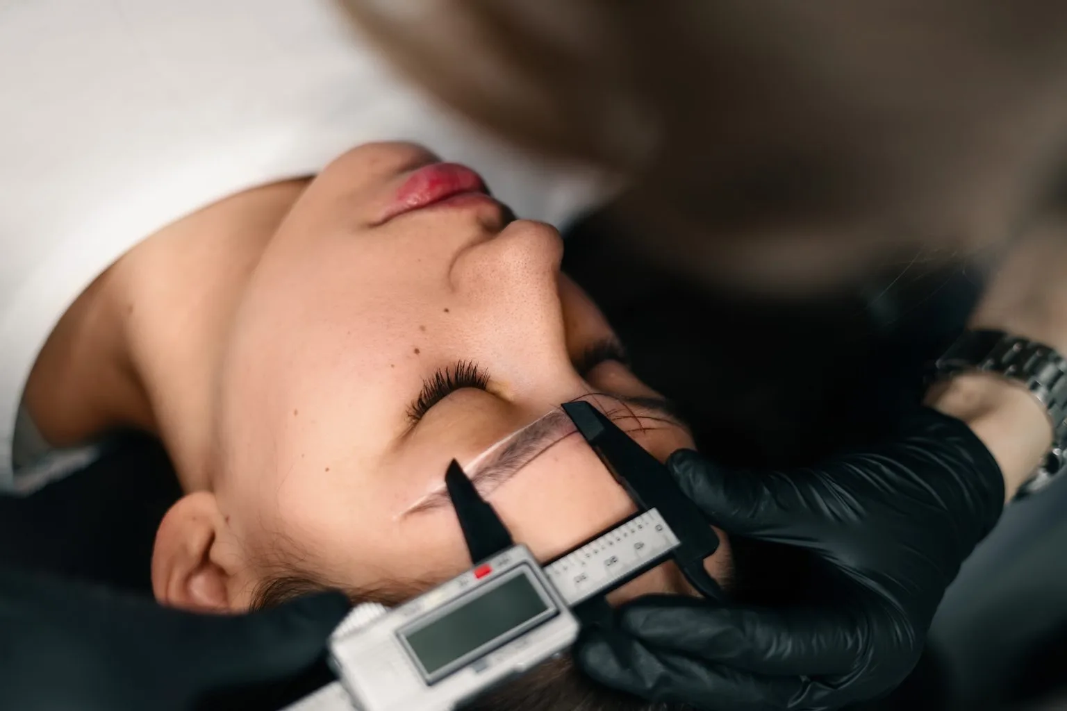 Precise brow-mapping at the Alesia Si studio — digital calipers in gloved hands against a client's brow