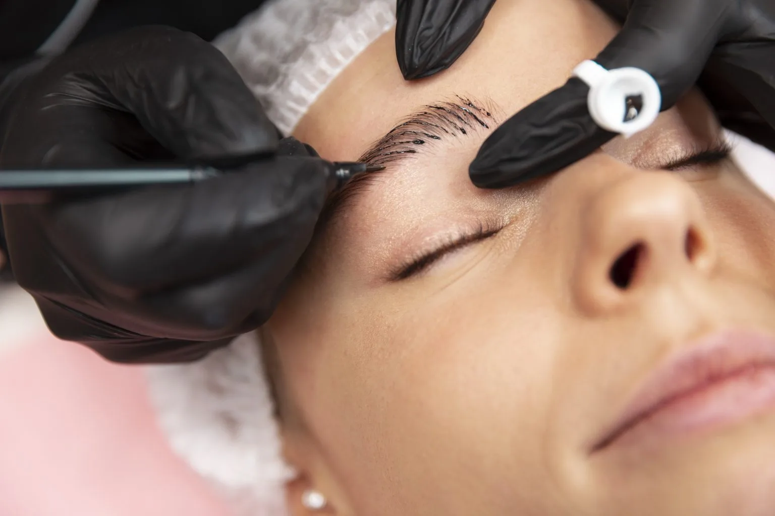 Microblading in progress — client reclined, gloved artist hands with hair net